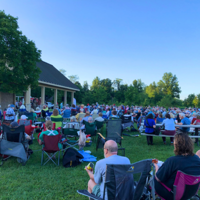 A crowd of people with camp chairs is gathered around the picnic shelter for a concert.